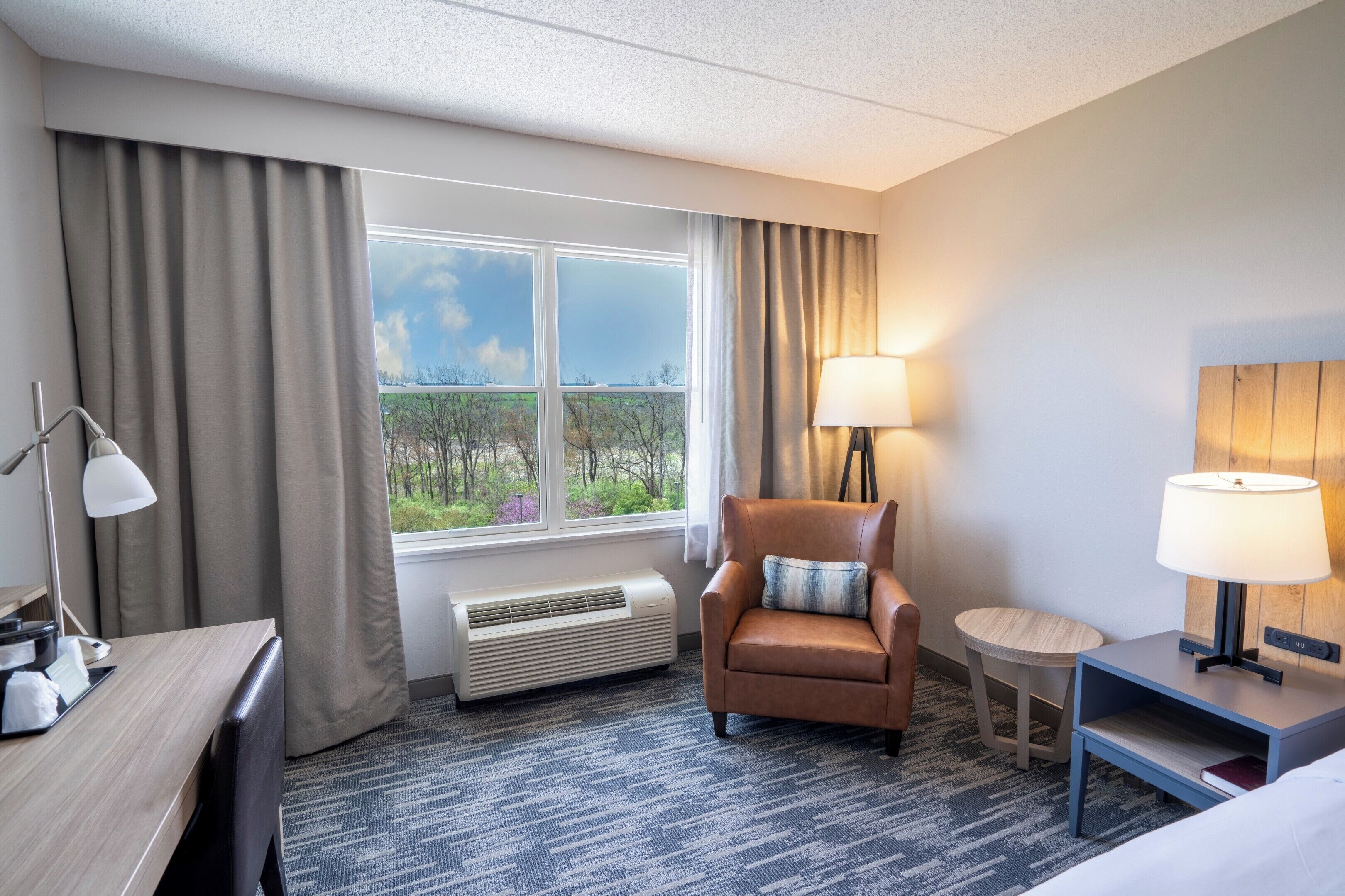 A hotel room with a bed, a desk, a chair, a small table, a lamp, a heater, and a window with curtains.
