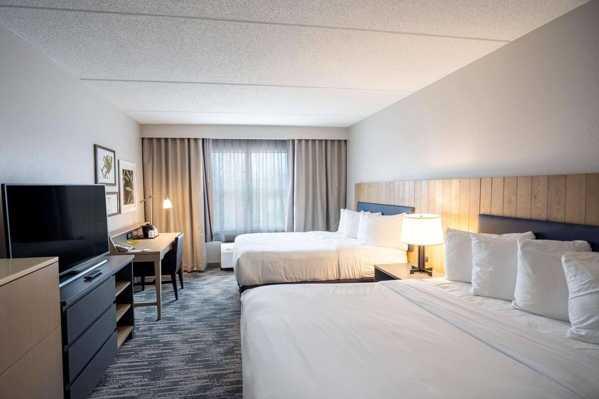 A hotel room with two beds, a desk, a television, and a window with curtains.