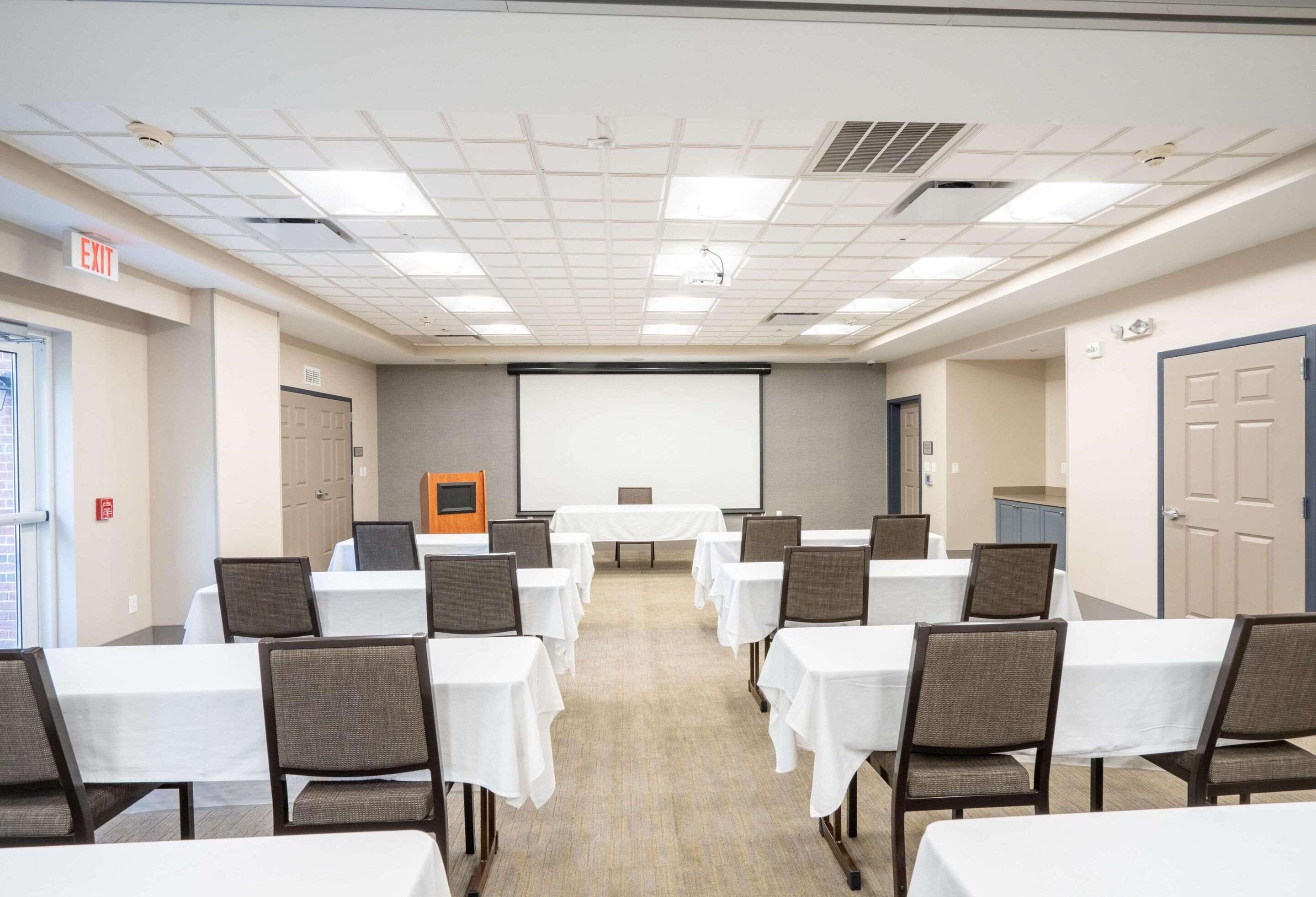 A conference room with tables and chairs, a projector screen, and an exit sign.