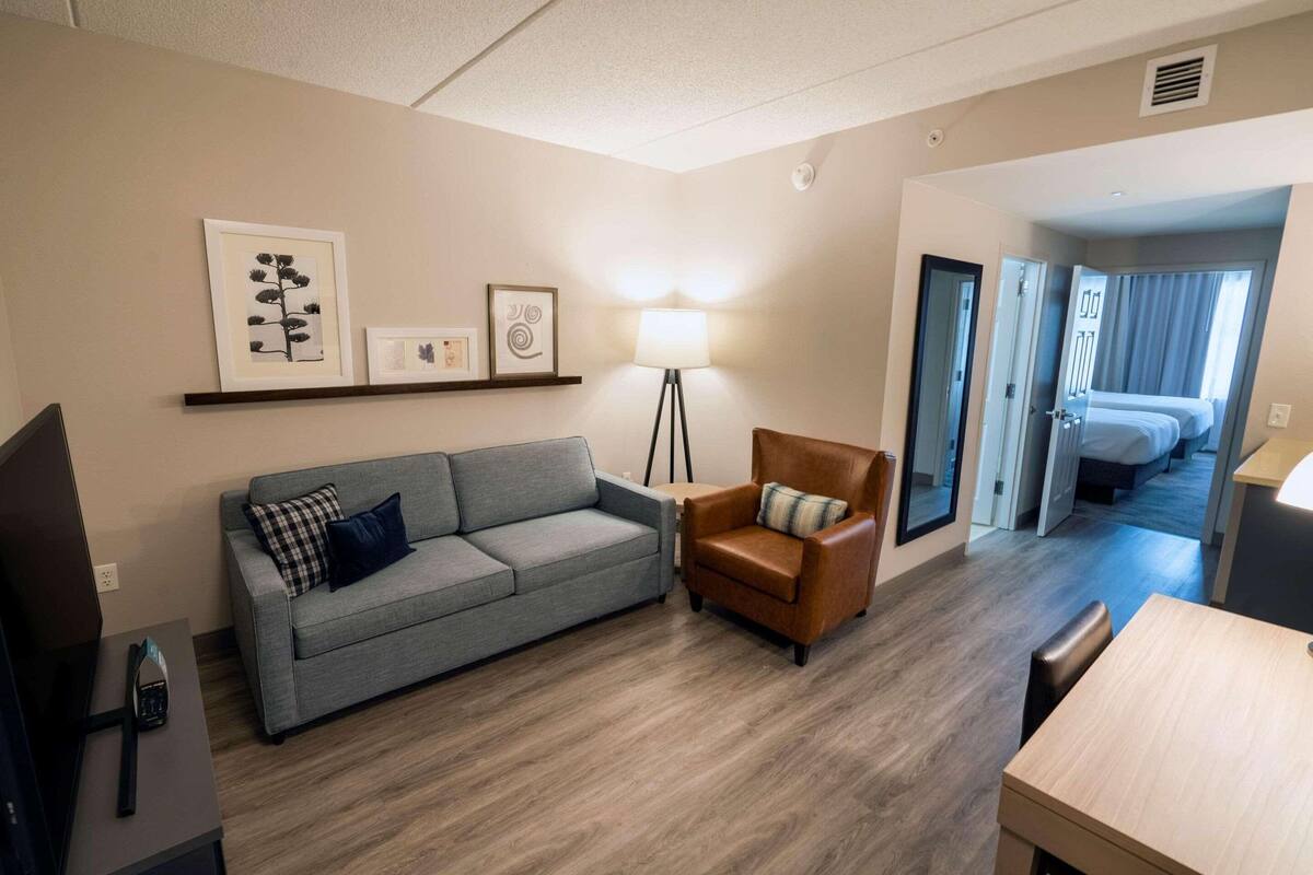 A modern hotel room with a grey sofa, a brown armchair, a desk, and a bed in the background.