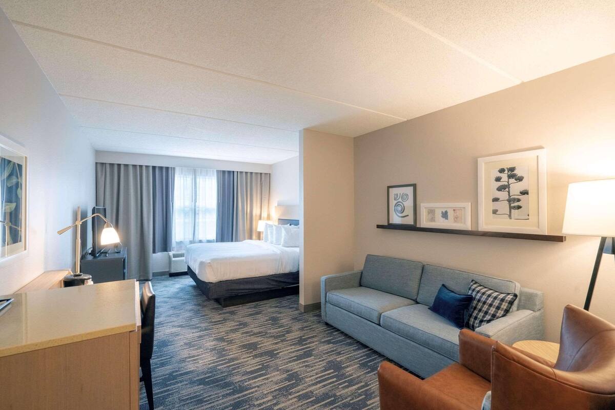 A hotel room with a bed, a desk, a sofa, and a chair. There are framed pictures on the wall and a lamp on the desk.