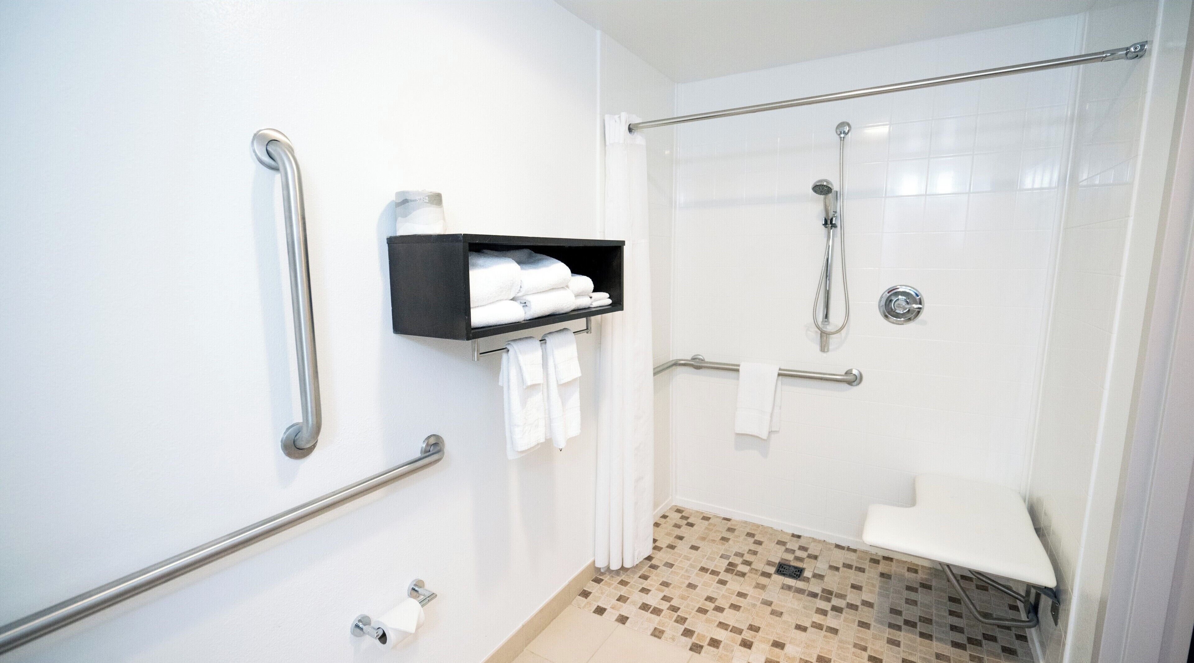 Un baño con ducha, barras de apoyo, estante con toallas y porta papel higiénico.