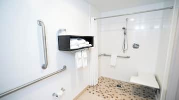 Un baño con ducha, barras de apoyo, estante con toallas y porta papel higiénico.