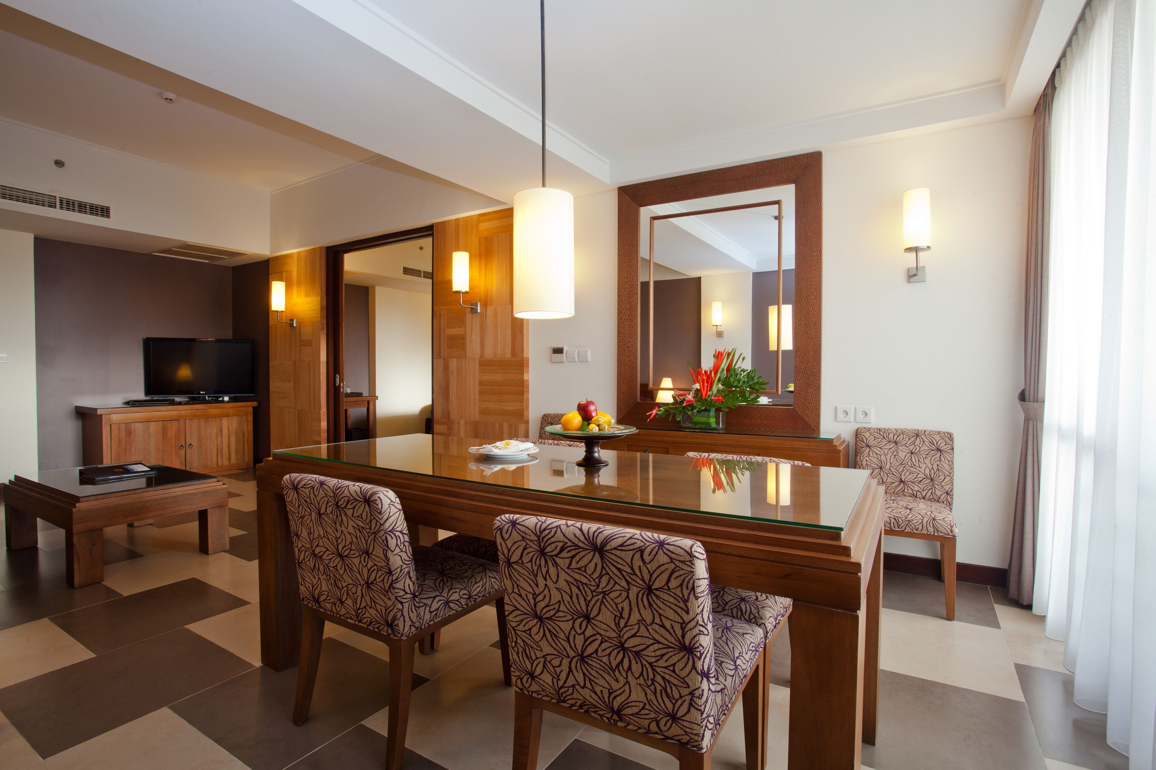 A hotel dining area with a glass-top table, patterned chairs, a television, and a mirror.