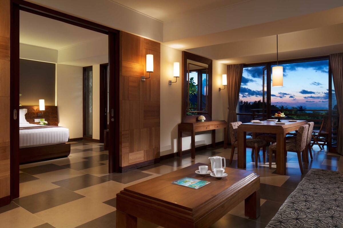 A hotel room with a bed, a dining area with a table and chairs, a coffee table, and a view of the sky through large windows.