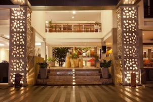 A hotel lobby with a central staircase, decorative pillars, and a balcony with plants.