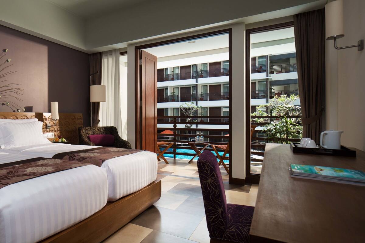 A hotel room with a large bed, a decorative wall piece, a balcony with a pool, and a view of a building.