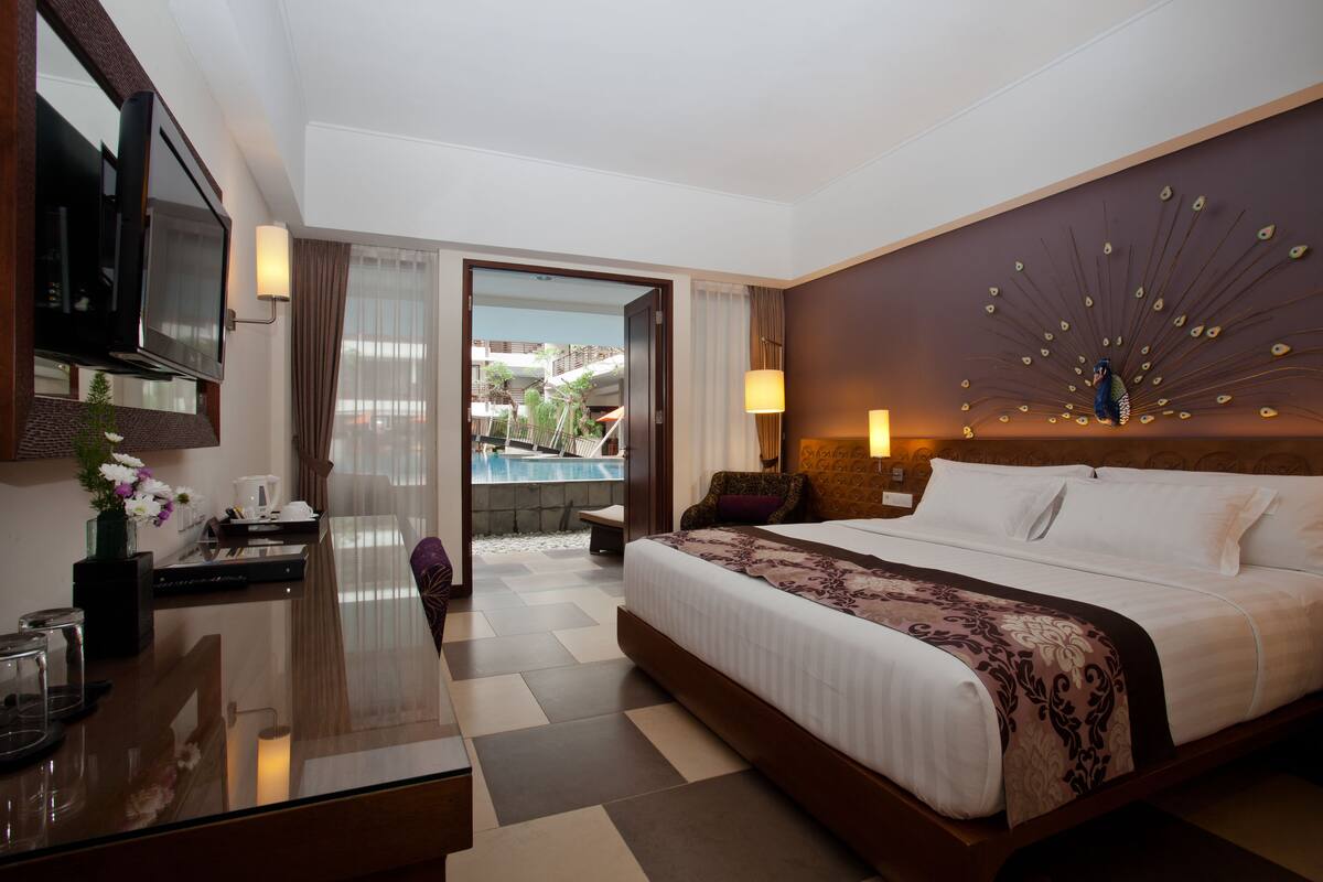 A hotel room with a large bed, a desk with a TV, a chair, and a view of a pool and outdoor area.