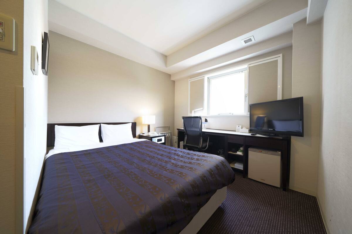 A hotel room with a bed, a desk with a television, a chair, and a small refrigerator.
