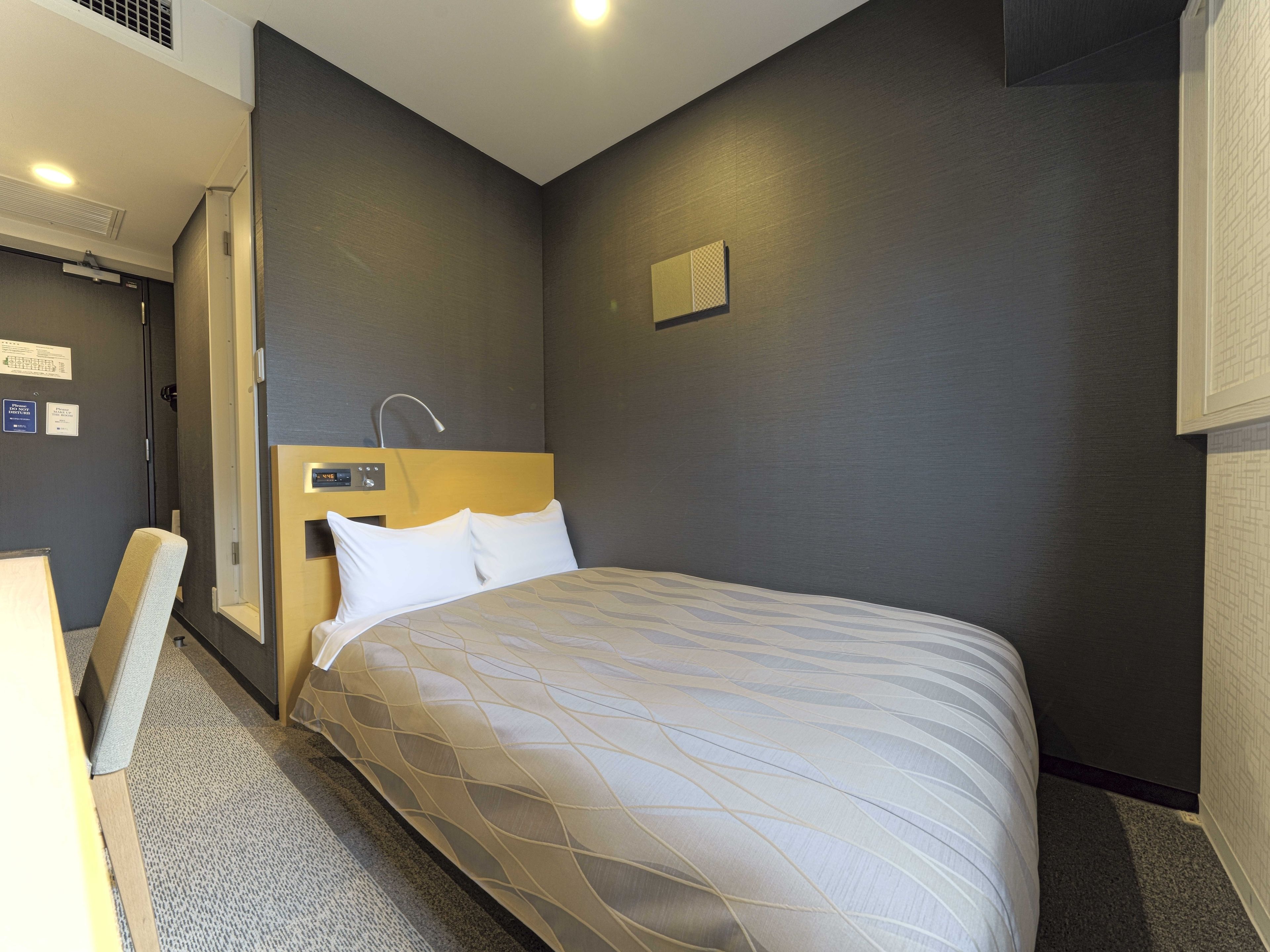 A hotel room with a bed, a headboard with a control panel, a shower area, and a small window.