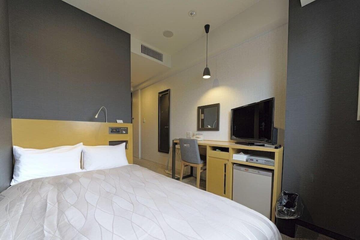 Quarto de hotel com cama, escrivaninha, cadeira, televisão e um pequeno frigorífico.