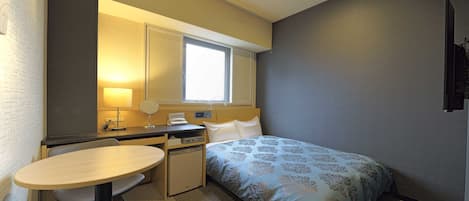 Habitación de hotel compacta con cama, escritorio, silla, mesita, espejo, ventana y televisor montado en la pared.
