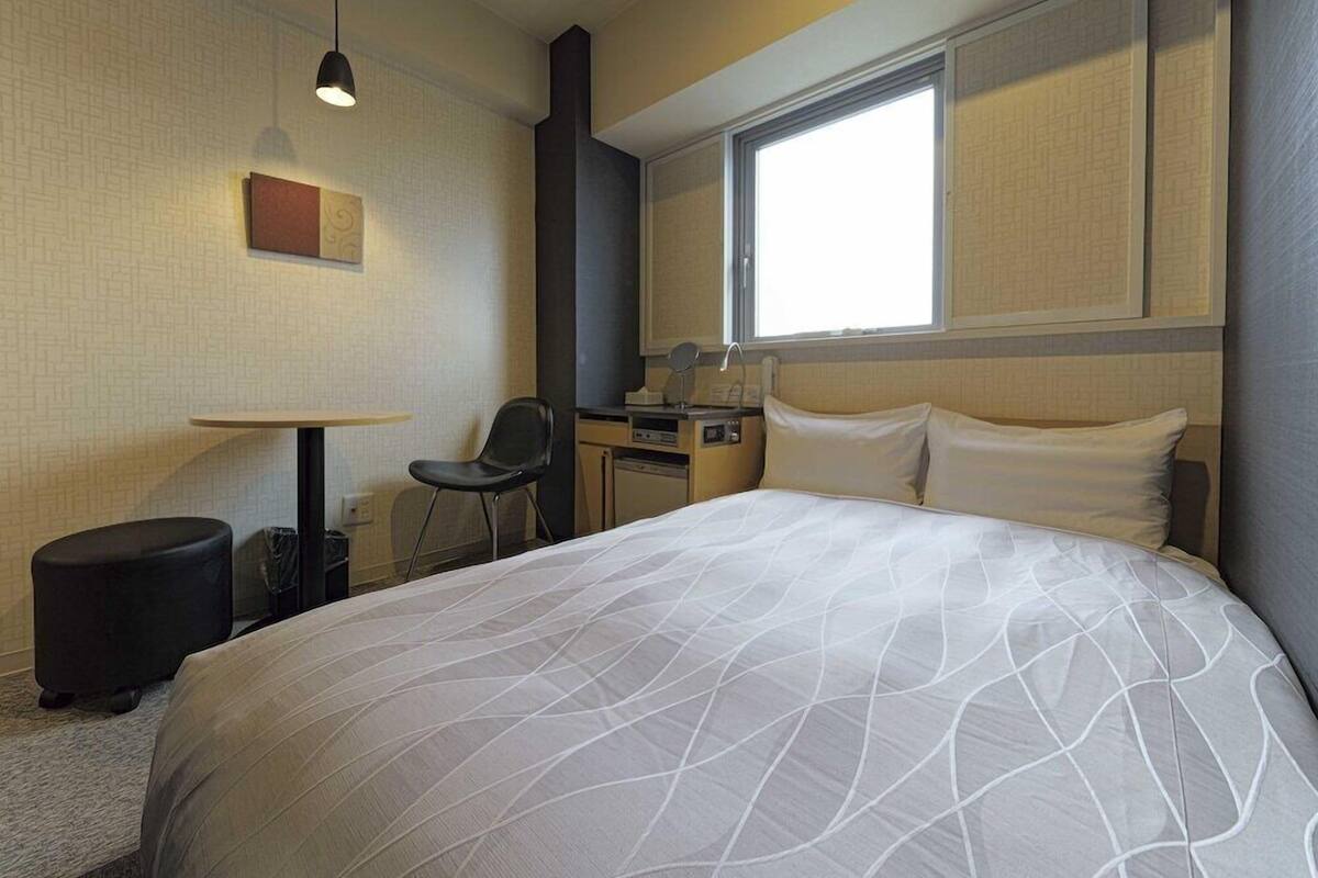 Quarto de hotel com cama, escrivaninha, cadeira e janela.