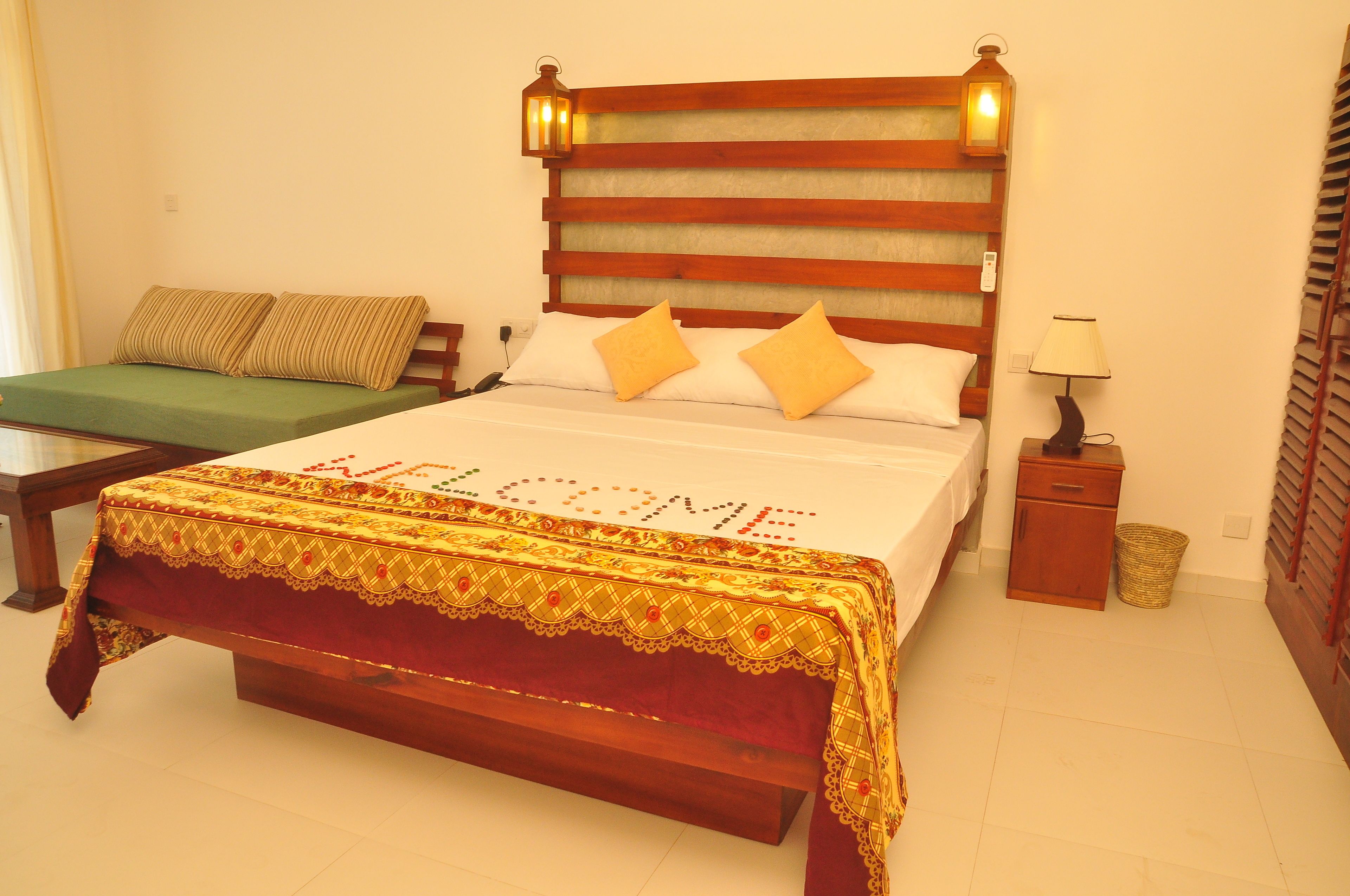 A bed with a 'WELCOME' sign, a bedside table, a lamp, and a small wooden cabinet.
