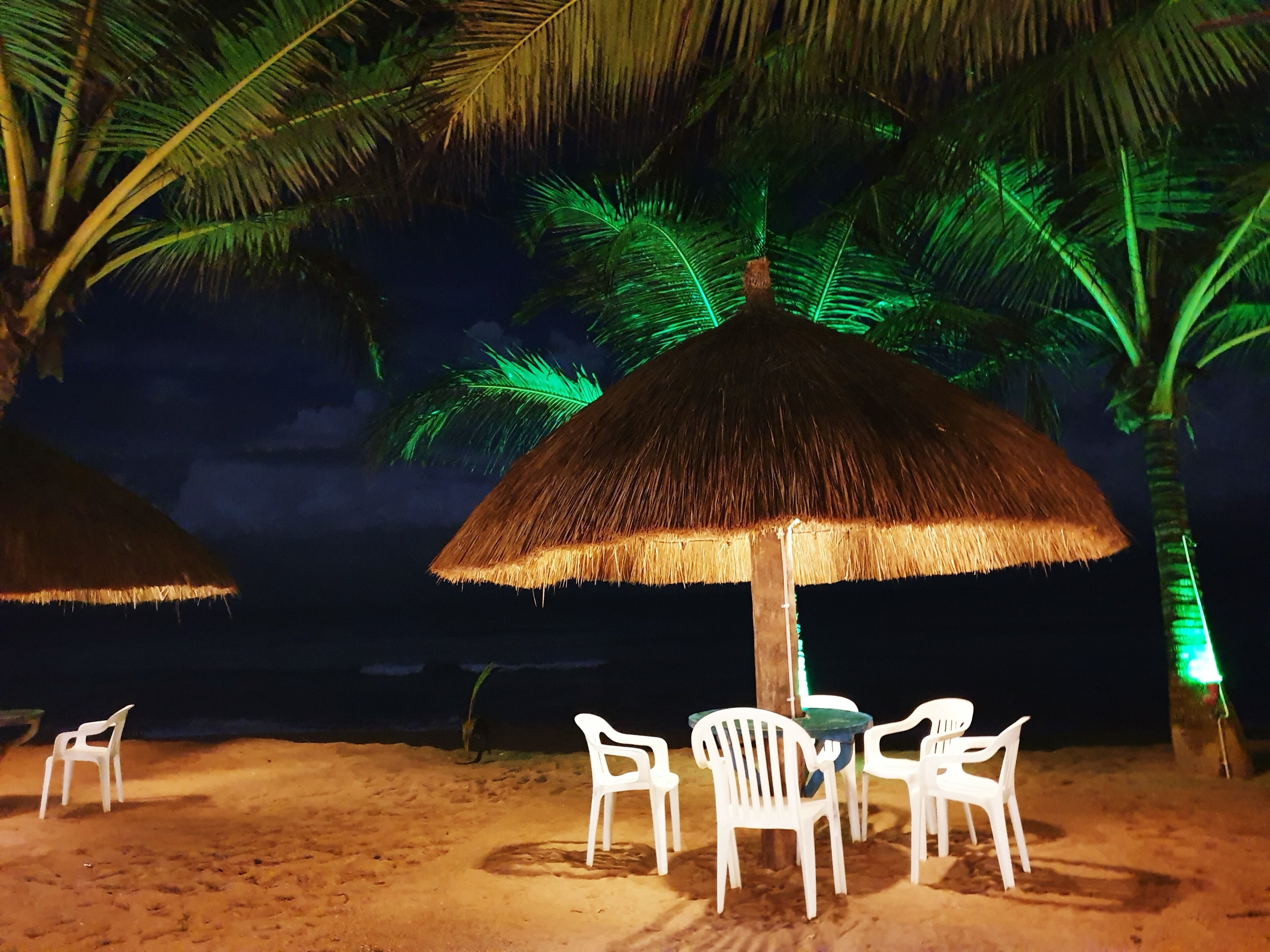 A beach setting with a thatched umbrella and white plastic chairs under it.