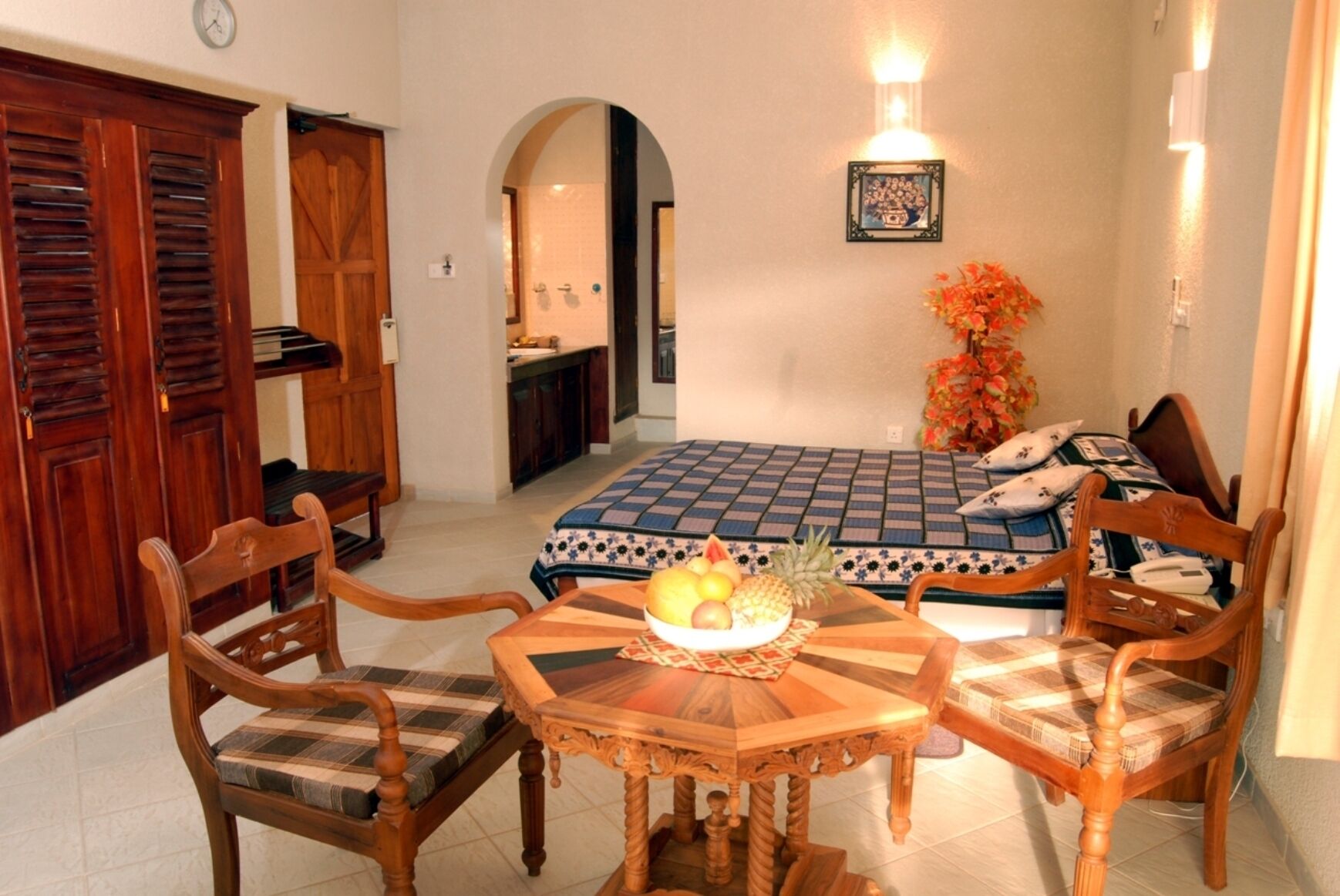 A cozy room with a dining table, chairs, a bed with a patterned bedspread, a small kitchen area, and a wooden wardrobe.