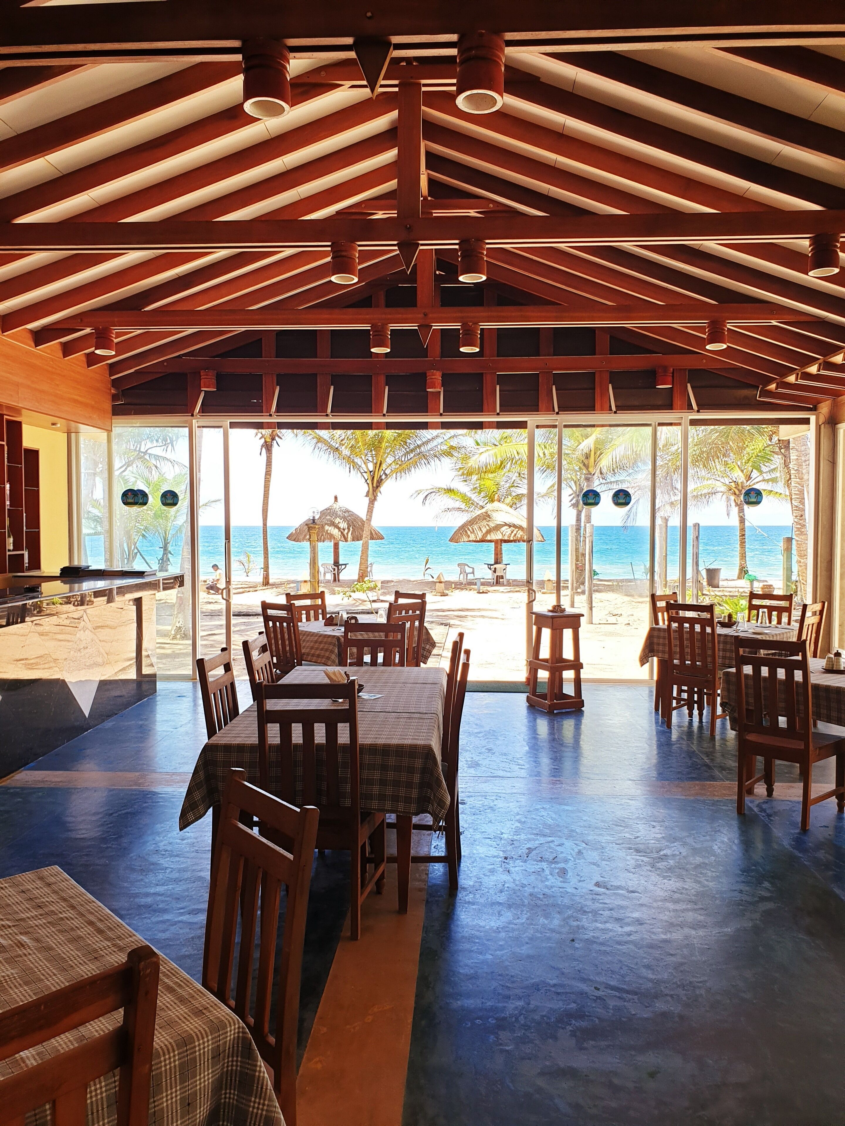 Un comedor interior con mesas y sillas de madera, amplios ventanales con vista a la playa y al océano.