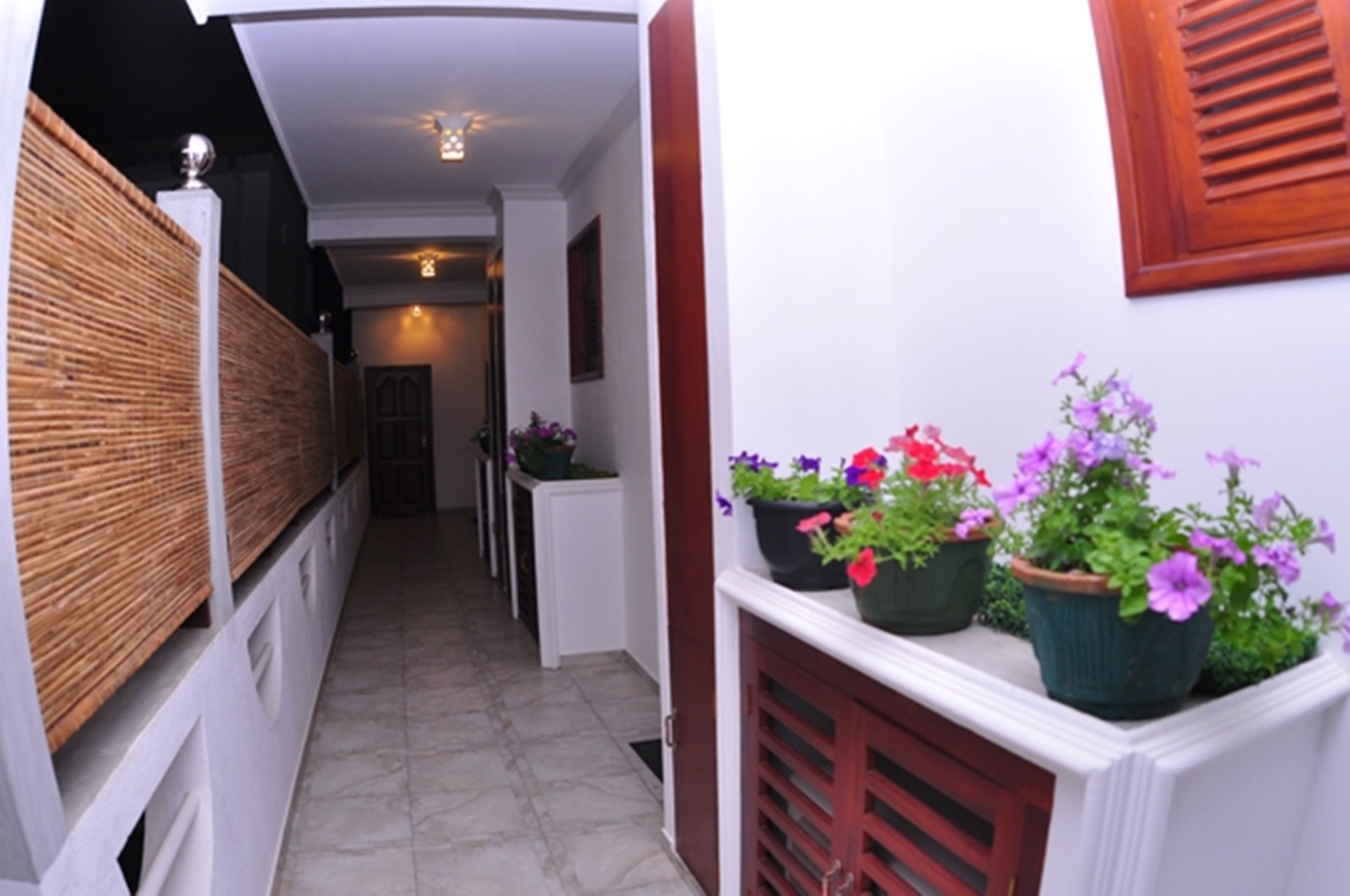 A narrow hallway with a tiled floor, white walls, and a wooden fence on the left. There are potted plants with flowers on the right.