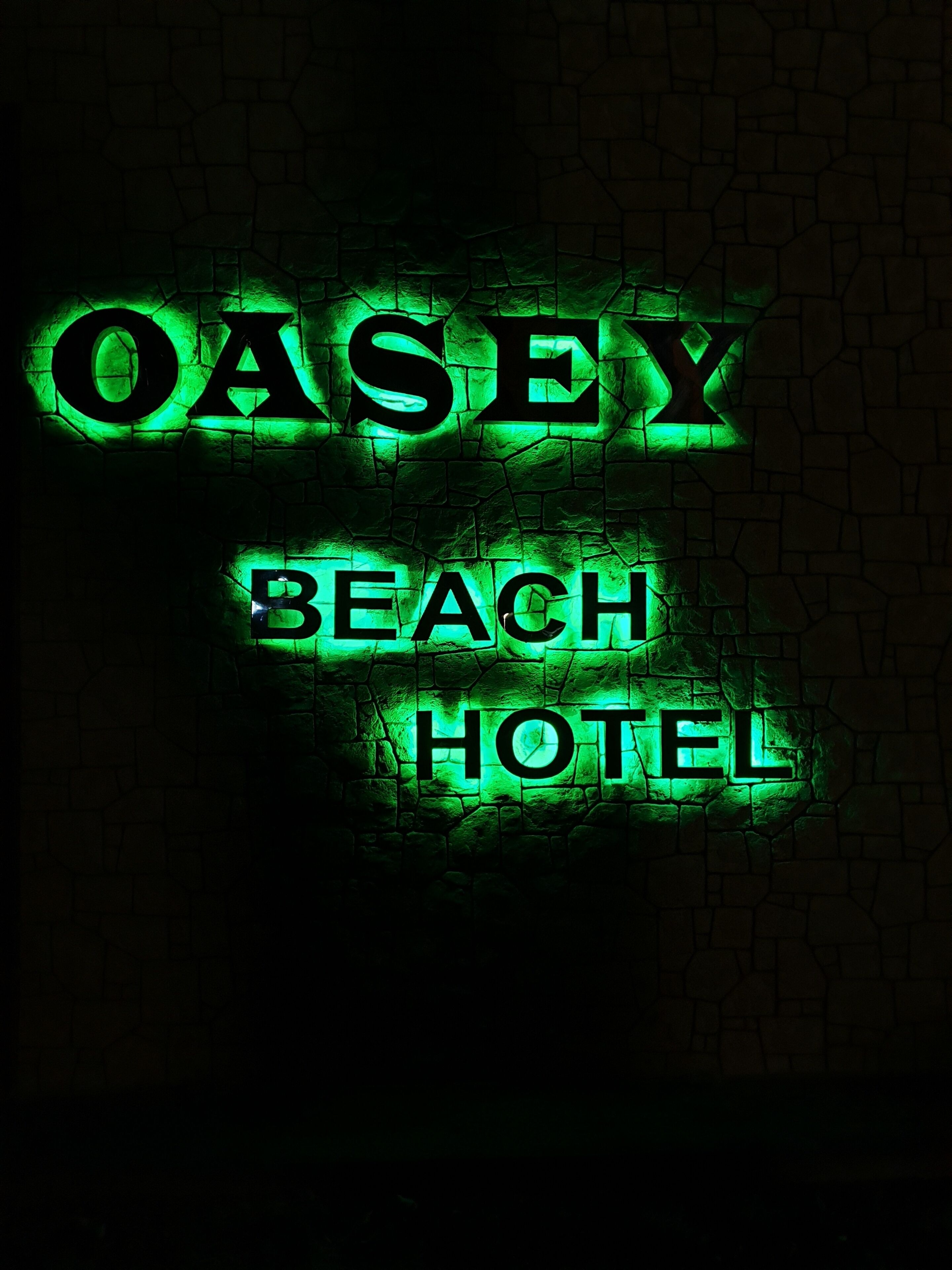 Une enseigne au néon avec l’inscription « OASEY BEACH HOTEL » sur fond de mur en pierre.