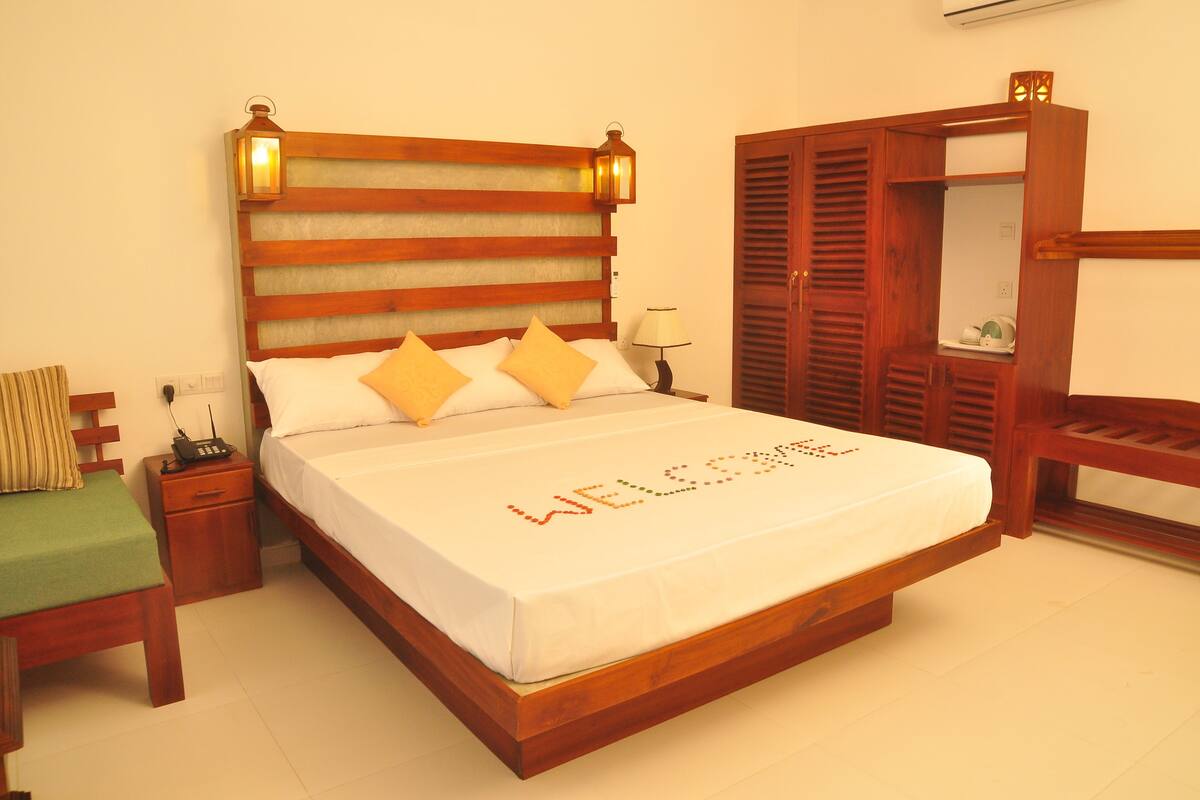 Una cama bien hecha con un cartel que dice 'WELCOME', una mesita de noche con lámpara, un armario de madera y una silla verde.
