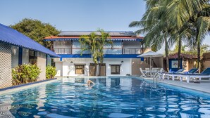 A swimming pool with a person swimming, surrounded by a building with a blue roof and white walls, and palm trees.
