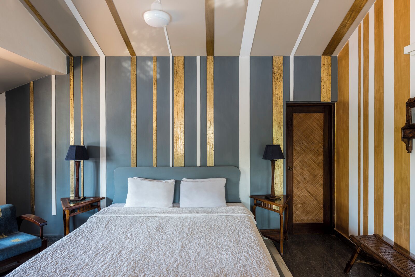 A hotel room with a large bed, two bedside tables with lamps, a blue armchair, and a door.