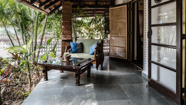 A covered patio area with a wooden ceiling, a glass table, a sofa with cushions, and a view of greenery outside.