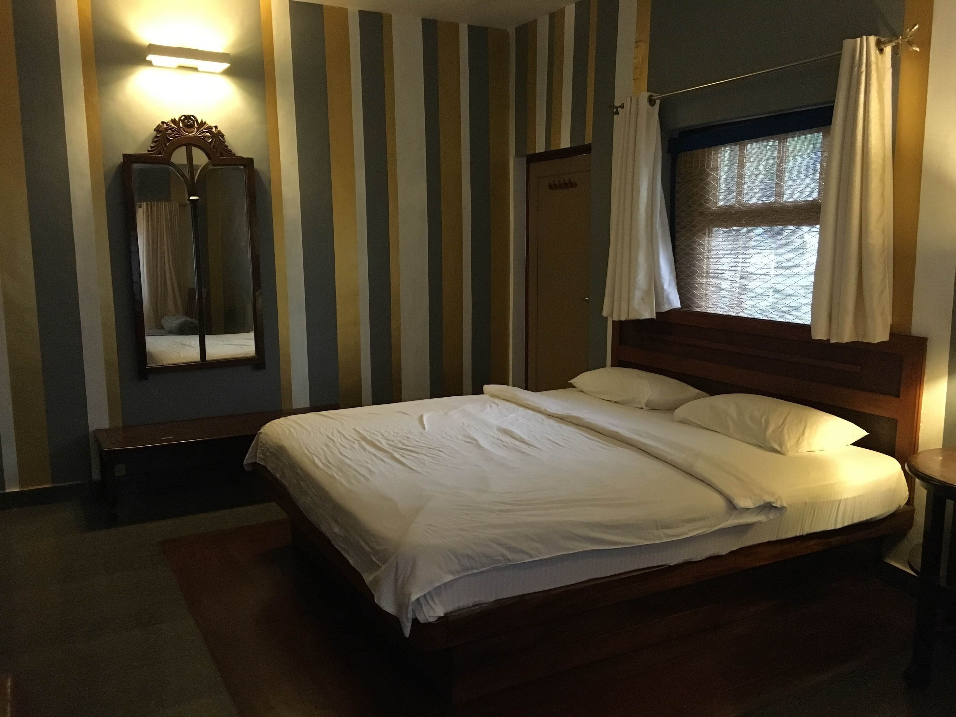 A hotel room with a bed, a mirror, a window with curtains, a wooden headboard, and a wall with vertical stripes.