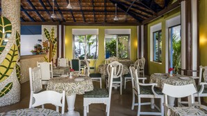 A restaurant with a thatched roof, white chairs, and tables covered with patterned tablecloths.