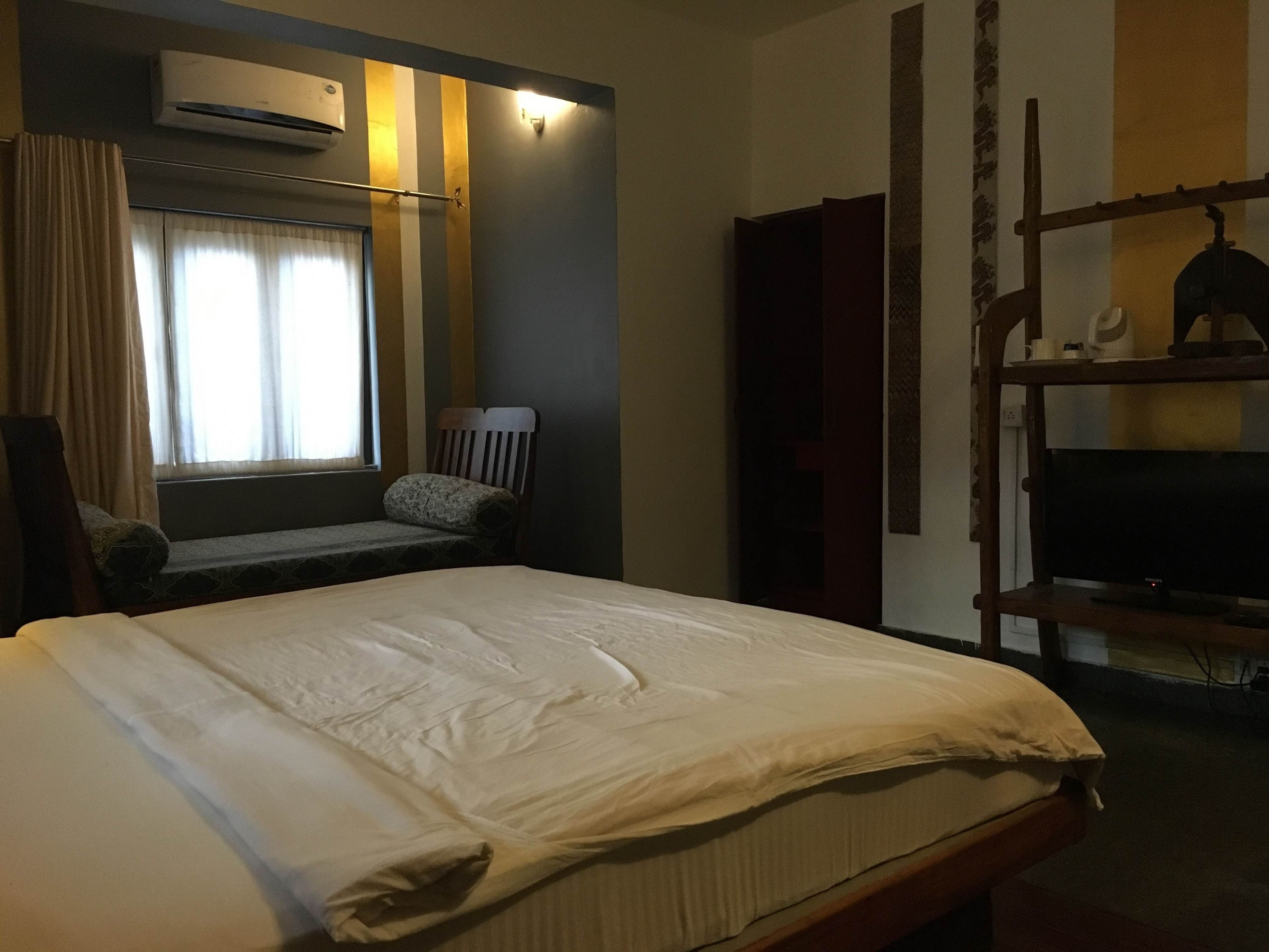 Une chambre d’hôtel avec un lit, une chaise, un téléviseur posé sur un meuble en bois, un appareil de climatisation et une fenêtre avec des rideaux.