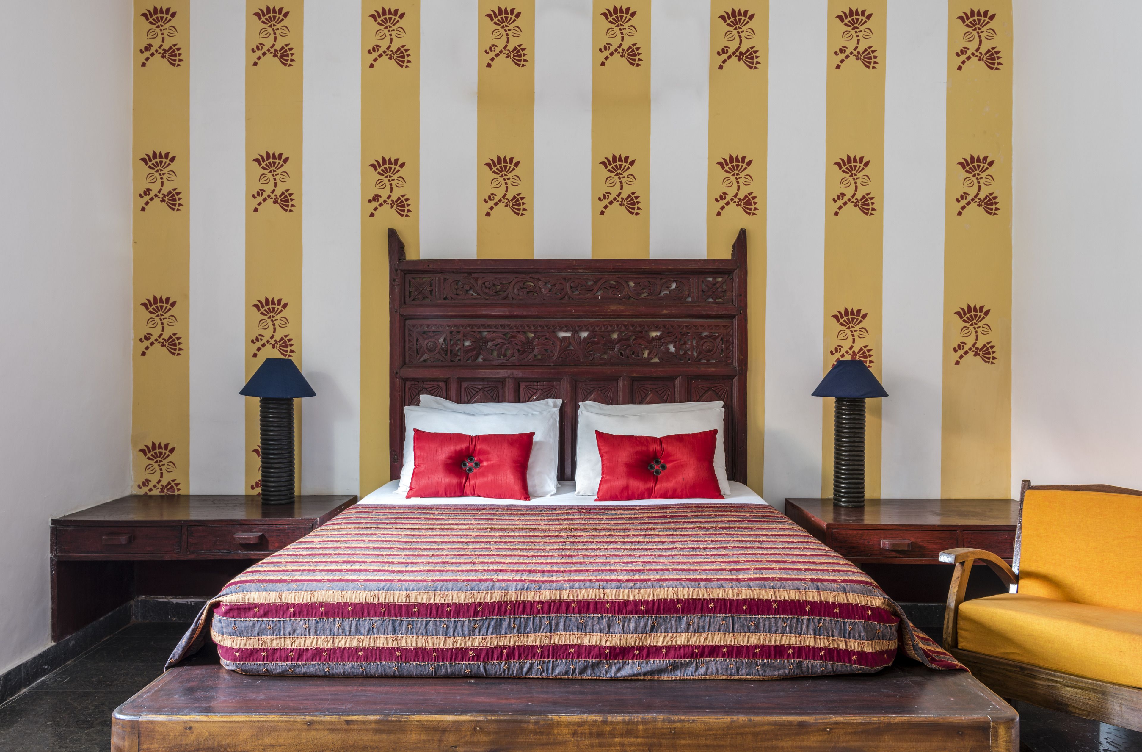 Una cama con cubrecama a rayas, dos almohadas rojas y un cabecero de madera. Hay dos mesitas de noche con lámparas y una silla amarilla.