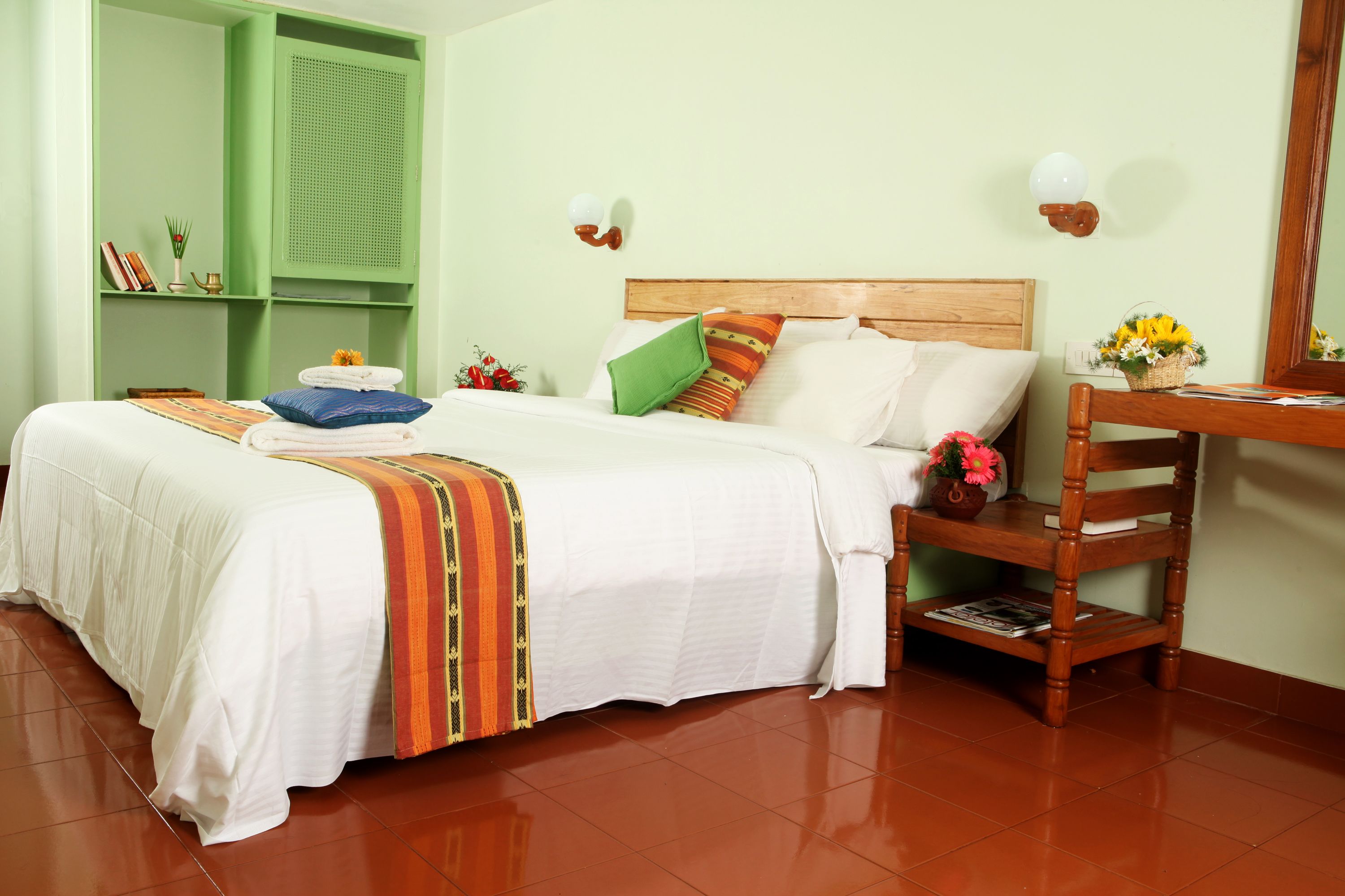 A neatly arranged bedroom with a large bed, a wooden chair, a small table with a vase, and a green shelving unit.