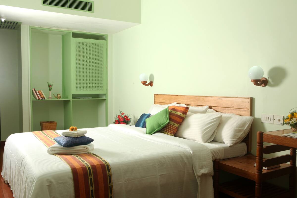 Een net opgemaakt bed met een gestreept dekbed, een houten hoofdbord en meerdere kussens. Er is een groene garderobe met spiegel en planken, een houten nachtkastje met een lamp, en een klein tafeltje met een bloemenvaas.
