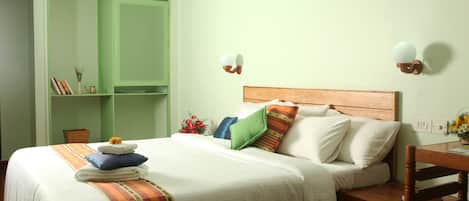 Una cama bien tendida con una colcha a rayas, un cabecero de madera y varias almohadas. Se observa un armario verde con espejo y estantes, una mesita de noche con lámpara, y una pequeña mesa con un jarrón de flores.
