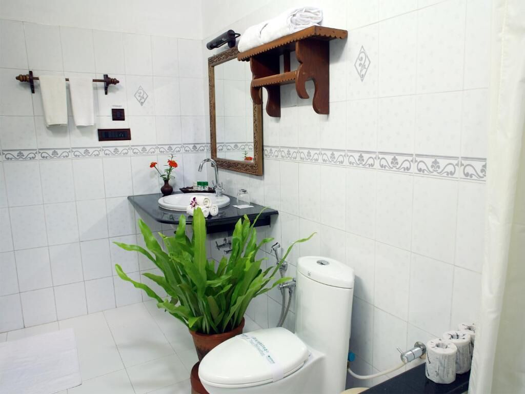 Un baño con paredes de azulejos blancos, un estante de madera con una toalla, un espejo, un lavabo, un inodoro, una planta en maceta y papel higiénico.
