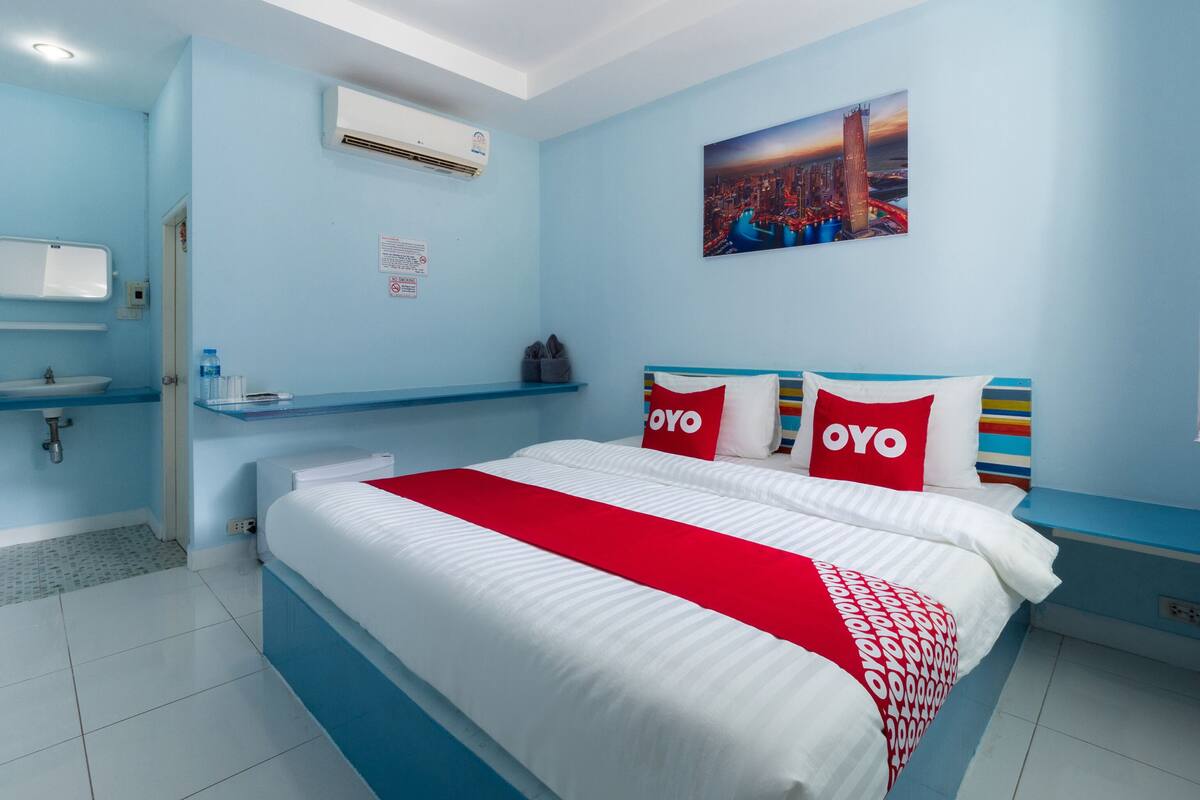 Una habitación de hotel moderna con una cama grande, un baño con lavamanos y espejo, y un cuadro de un paisaje urbano en la pared.