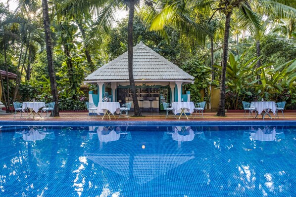 Un espace aménagé au bord de la piscine, comprenant un kiosque, des tables et des chaises, le tout entouré d’une végétation luxuriante.