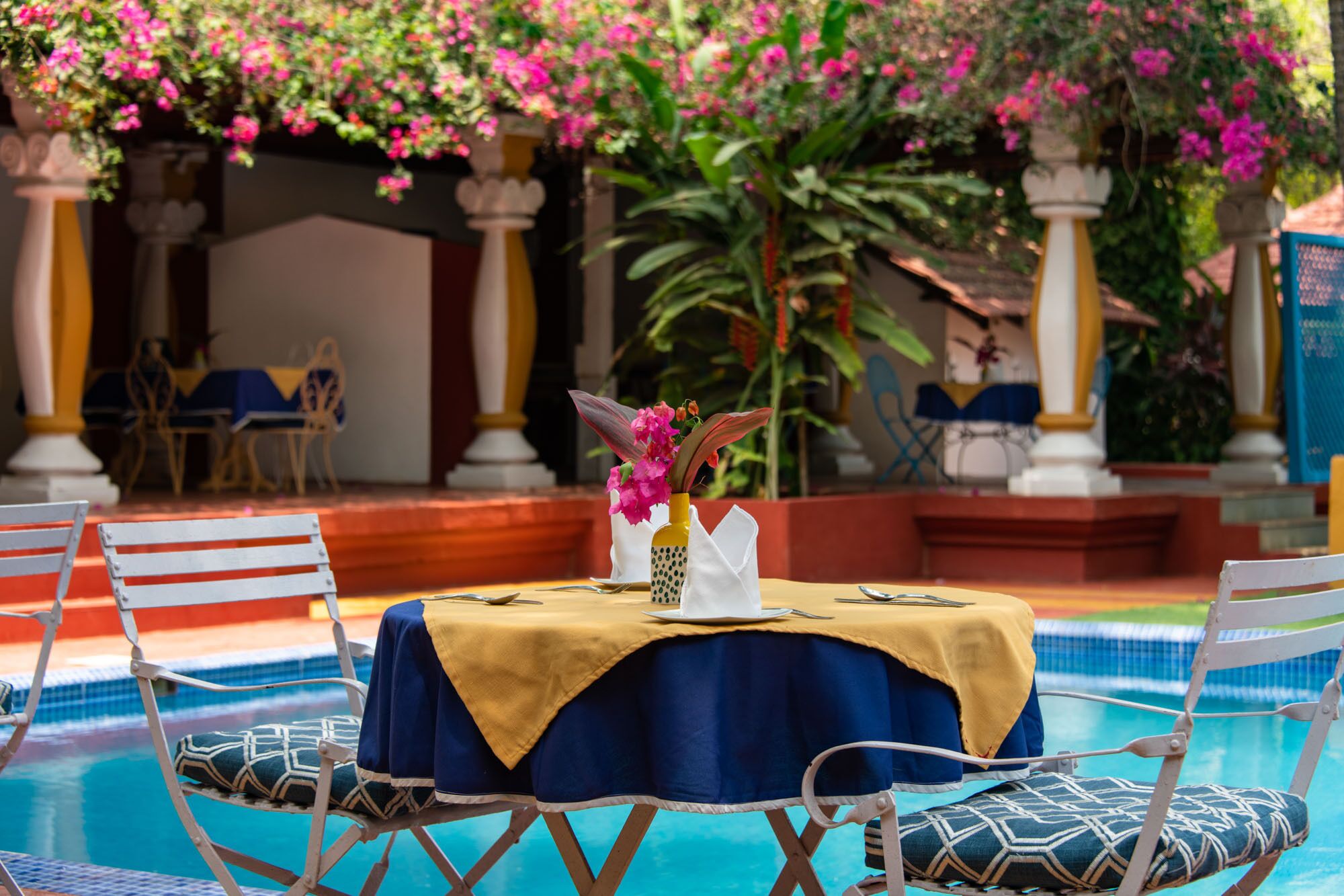 Un espace repas aménagé au bord de la piscine, avec une table dressée pour deux personnes, entourée de chaises dotées de coussins à motifs.