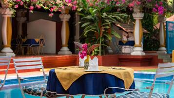 Un espace repas aménagé au bord de la piscine, avec une table dressée pour deux personnes, entourée de chaises dotées de coussins à motifs.
