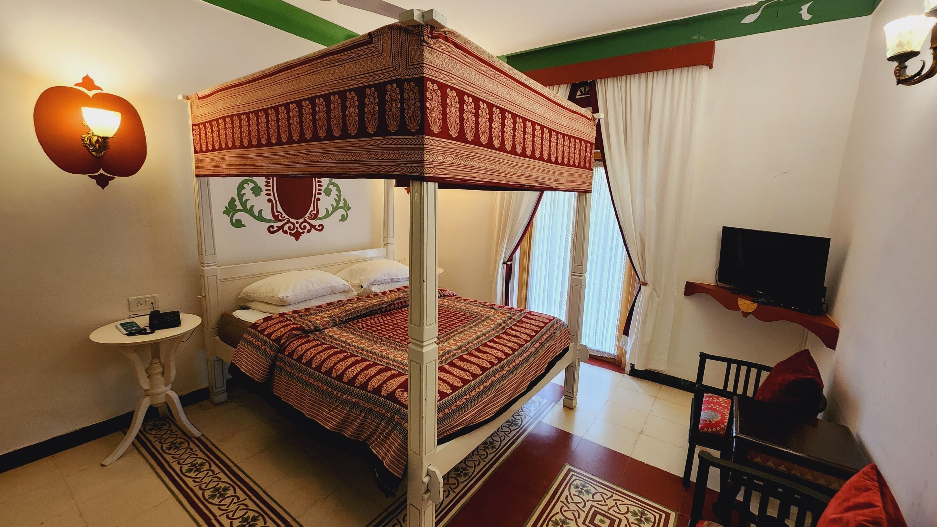 A four-poster bed with a patterned bedspread, a bedside table with a lamp, a television on a stand, and a chair with a cushion.
