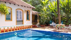 A swimming pool with blue tiles, a white building with yellow trim, and outdoor furniture. - Casa Anjuna (Anjuna)
