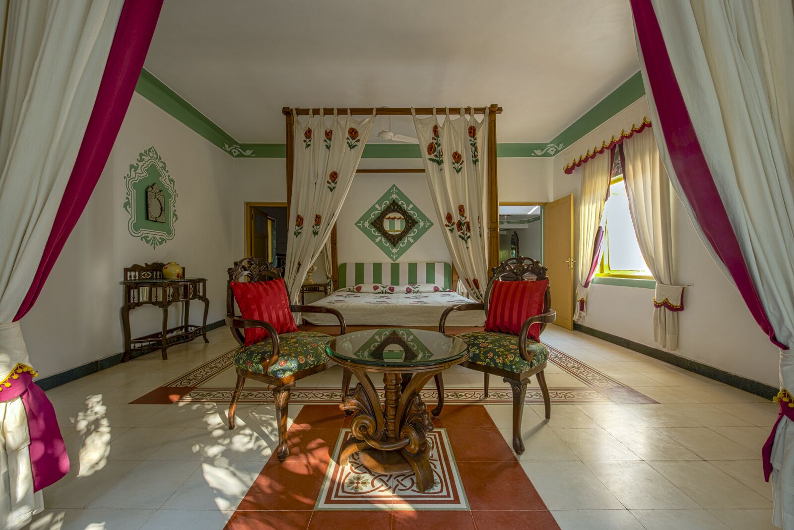 A four-poster bed with a canopy, two chairs with red cushions, a small table, and a decorative mirror on the wall.