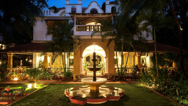 A well-lit courtyard with a central fountain, surrounded by palm trees and potted plants, leading to a building with a traditional architectural style. - Casa Anjuna (Anjuna)