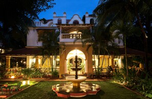 Un patio luminoso con una fuente central, rodeado de palmeras y plantas en macetas, que conduce a un edificio de estilo arquitectónico tradicional.
