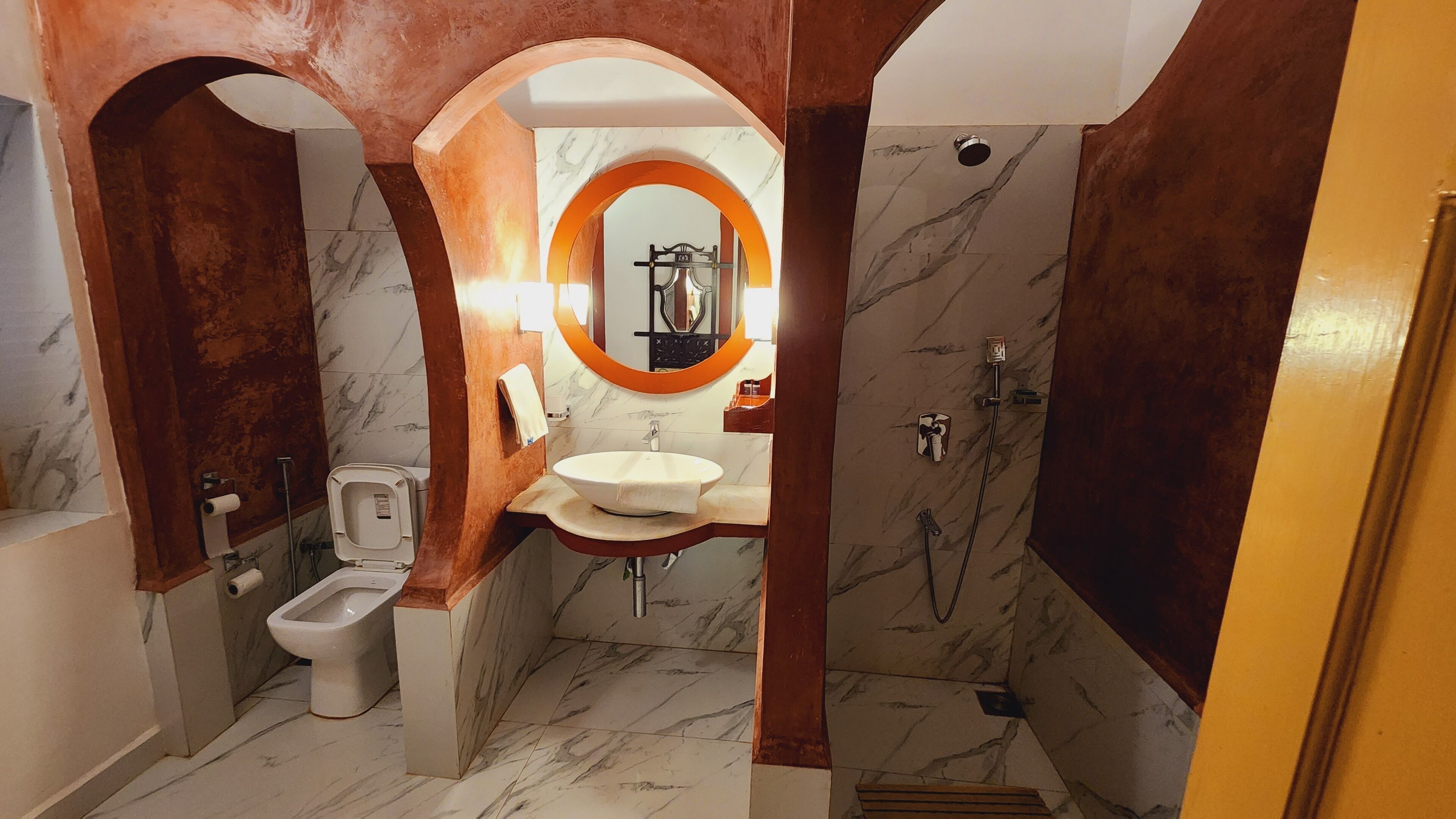A bathroom with a marble floor, a toilet, a sink, a shower, and a mirror with an orange frame.