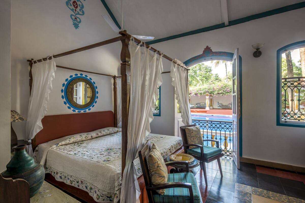 Uma cama com dossel e cortina, um espelho redondo, e vista para uma piscina e um jardim através de uma porta aberta.