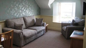 A cozy living room with a grey sofa, a wooden chair, and a small table. - Glenholme Apartments (Tenby)
