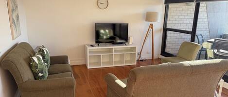 A living room with a TV on a white stand, two armchairs, a lamp, and a window with a view of a brick building.