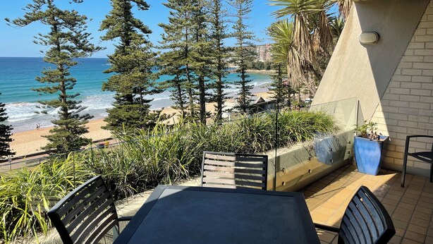 Un balcone con vista sulla spiaggia e l'oceano, dotato di tavolo e sedie, circondato da piante e alberi.