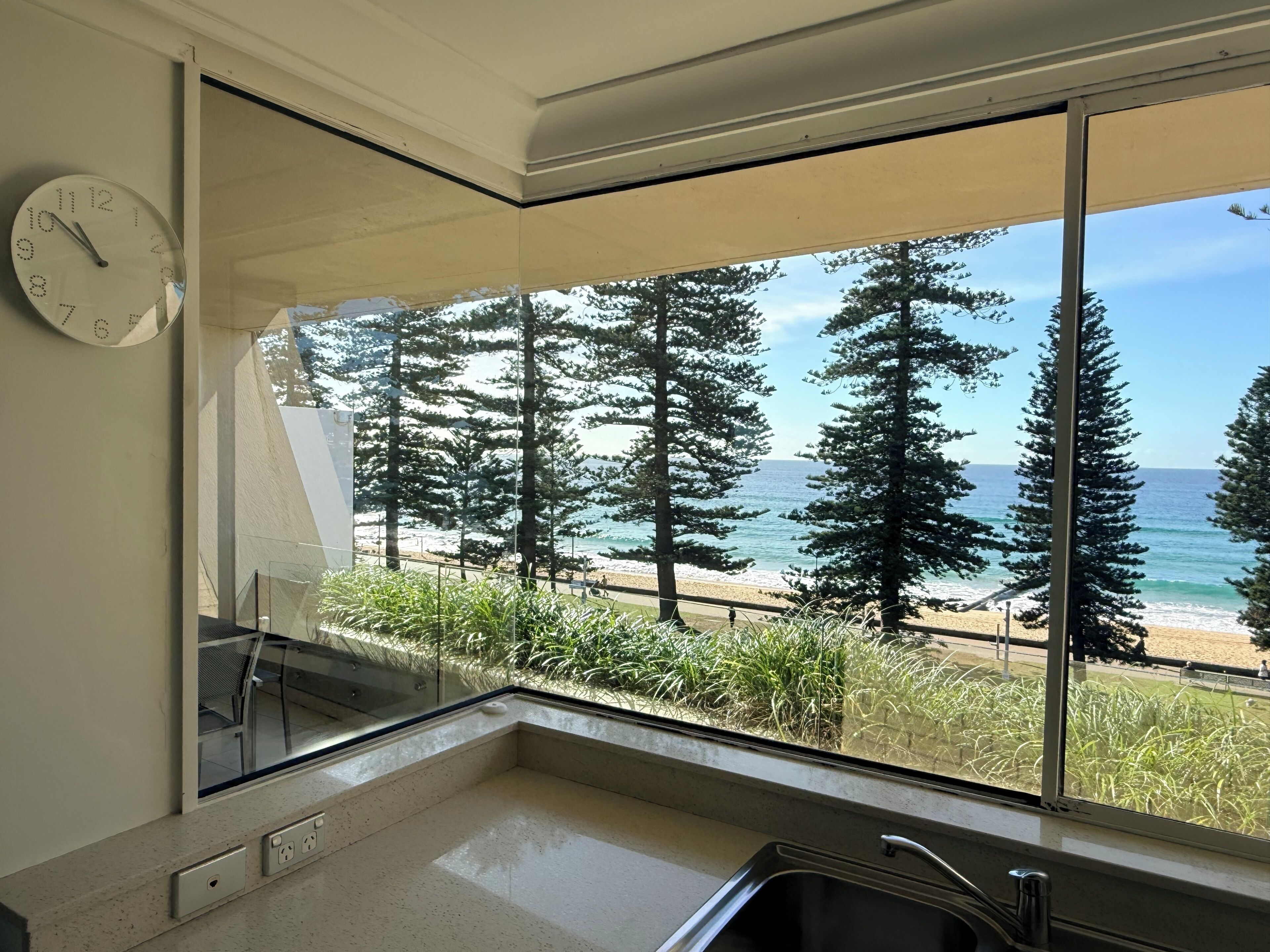 A kitchen with a large window overlooking a beach, trees, and a clock on the wall.